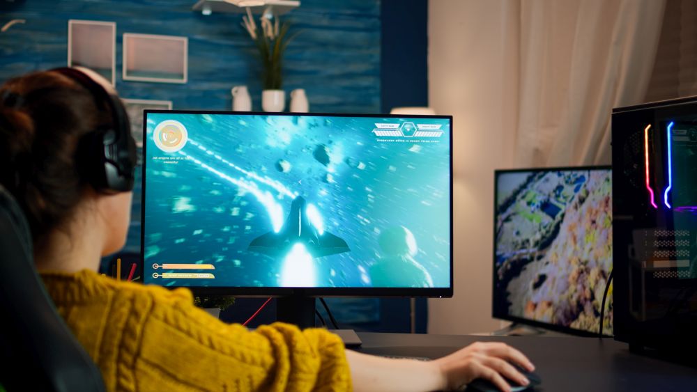 Osoba u žutom džemperu i sa slušalicama igra video-igru na velikom gaming monitoru, dok se pored nalaze dodatni ekran i računar sa RGB osvetljenjem